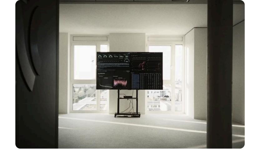 A large dashboard screen on a stand in an otherwise empty white office room, showing training metrics, charts and a results table.